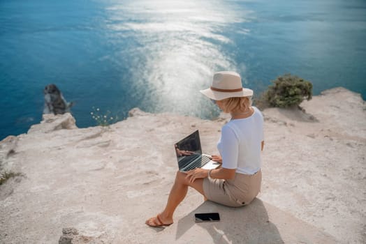 Freelance women sea working on the computer. Good looking middle aged woman typing on a laptop keyboard outdoors with a beautiful sea view. The concept of remote work