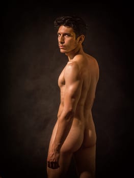 Portrait of naked handsome young man with languishing look covering crotch with his hands. In a studio with a dark backdrop, an athletic, handsome young man stands completely nude looking at camera.