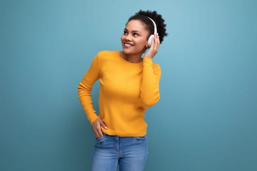 pretty positive20s latin woman with afro hair in casual yellow sweater listening to music in wireless headphones.