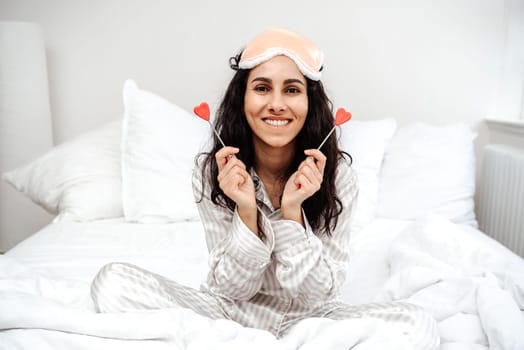 A young Arab woman wakes up in a romantic setting. The girl in anticipation of the holiday holds hearts in her hands. Valentine's Day. A young woman sits in a bright room in pajamas on a cozy bed