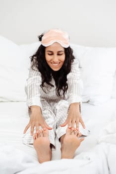 Charming cheerful young woman stretching legs and doing yoga in her bed. Waking up concept. Girl stretches her hands to her feet. Feet in focus and in the foreground girl in defocus in the background