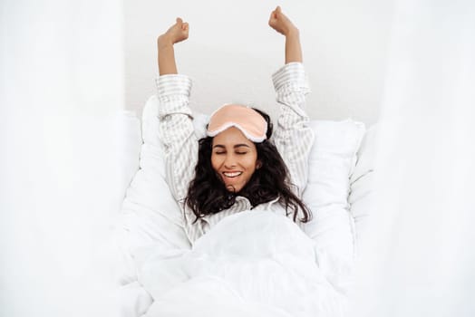 A woman lies in bed and pulls her hands up after waking up. Healthy lifestyle. Wellness concept. A young beautiful brunette woman wakes up in her bed fully rested.