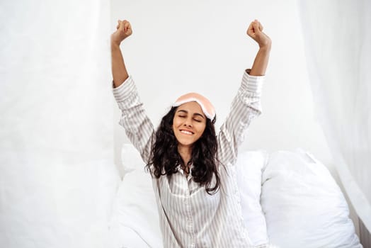Young Arab woman has just woken up in her snow-white bed. Girl stretches her hands up having fun. Stretching in the morning, we start our day with positive emotions. Girl closed eyes with happiness
