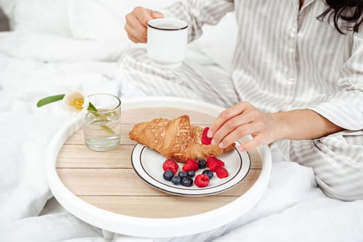 The best start to the day is breakfast in bed. Girl's hands take a berry and a cup of coffee. Close up of croissant with berries and flower. Detail of a girl in pajamas sitting on a white bed.