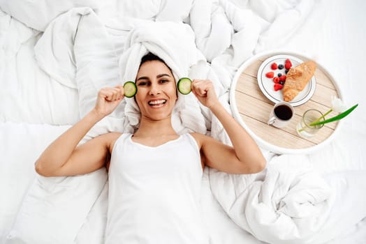 Young woman taking care of their skin at home. She lies on the bed after a shower and is ready to make a cucumber mask. On a snow-white bed there is a tray with breakfast, a cup of coffee and a flower