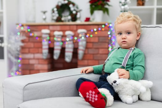 A little boy with curly blond hair sits on a gray couch. The child holds a white cuddly toy in his hand. A brick fireplace can be seen in the background. The fireplace is decorated with colorful Christmas lights.