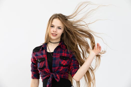 A girl in a youthful outfit poses for a photo against a white background. She is combing her fingers through her long, flowing hair. The woman is wearing a red and blue checked shirt.