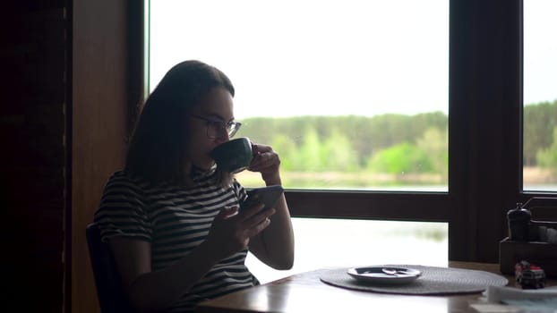 A young woman sits in a cafe by the window and chats on her smartphone. A girl with a phone in her hands at a panoramic window overlooking the river. 4k