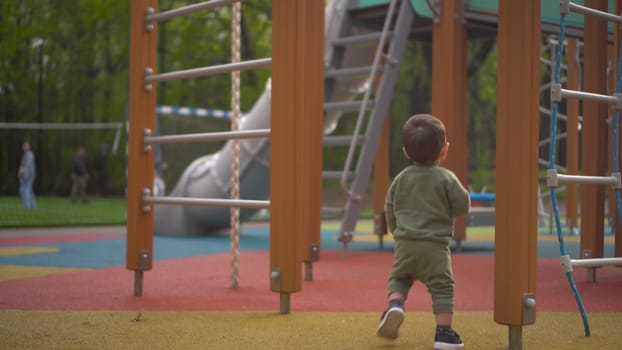 A two-year-old child walks on a playground among slides and swings. Little son in the park. 4k