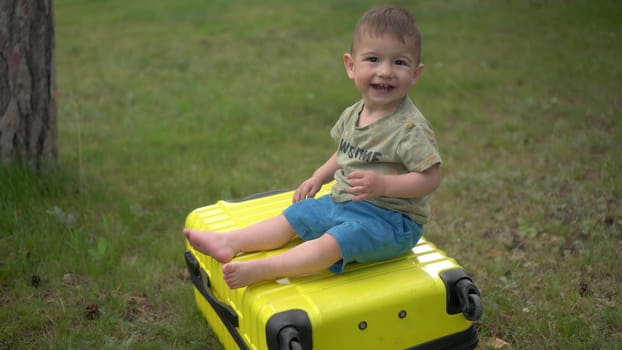 A little boy sits on a yellow suitcase in nature. A one-year-old child sits on a suitcase and rolls down. 4k