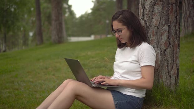 Young woman with laptop in nature. A girl with glasses sits under a tree and types on a laptop. Freelancer woman. Closeup. 4k