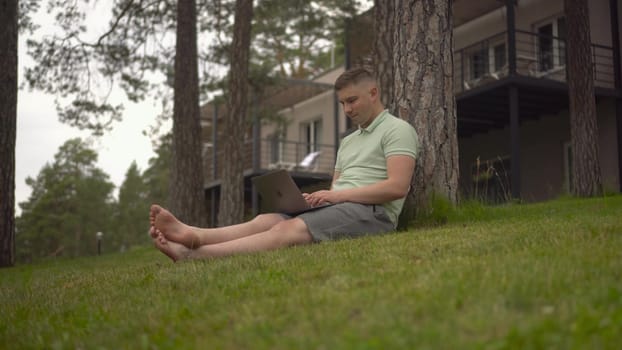 A young man sits under a tree and works on a laptop. A man with a laptop in nature sitting on the grass. 4k