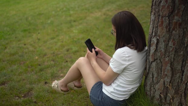 A young woman sits on the grass under a tree and chats on her smartphone. A girl with glasses is resting in the park with a phone. 4k