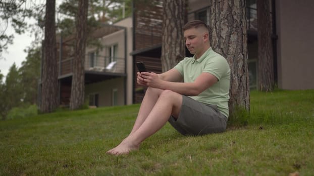 A young man sits under a tree near a country house with a phone in his hands. A man is texting on a smartphone in nature on the grass. 4k