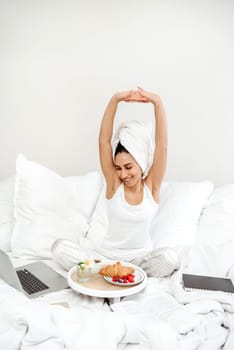 Happy woman stretching in bed in the morning with breakfast tray. The girl is ready to start work and perform tasks after a delicious healthy breakfast. A towel is wrapped around the woman's head.