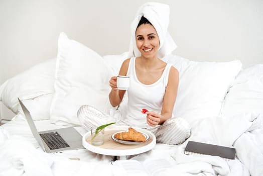 Young beautiful girl of Hispanic appearance sits on the bed in the morning after a shower Taking breakfast and starting a working day on a laptop. Girl holding a berry and a cup of coffee in her hands