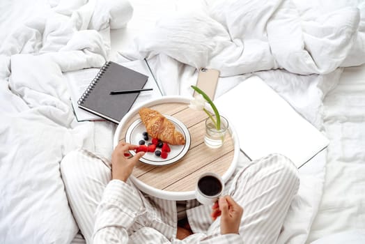 Healthy breakfast in bed croissant and fresh berries. The girl holds a cup of coffee in her hand and is ready to enjoy the meal. Starting a productive day in bed with work supplies laptop and notebook