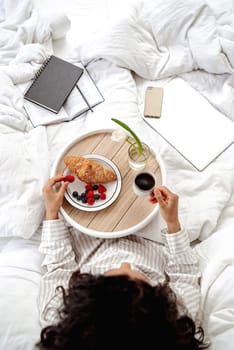Top view aesthetics of morning and breakfast in bed. Woman going to eat berry and croissant. The girl organized the workspace and starts her morning with a productive working day