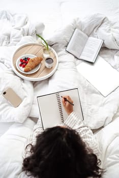 Cozy relaxation in bed. Top view of woman in white pajamas making note with breakfast on the bed. Woman daydreaming after reading a book and making notes for the future or wish list