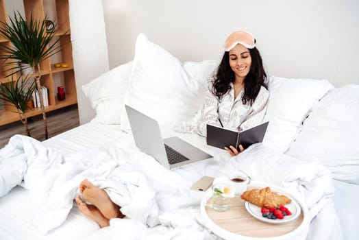 Young beautiful arab woman sitting on the bed making a note. Woman note tasks on laptop and writing down plans. A good start to the working day in a homely atmosphere over breakfast.