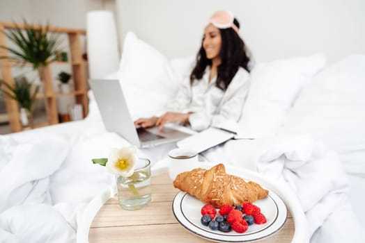 Woman working with laptop, eating fruit breakfast in bed at home or hotel in the morning. Girl wearing pajames on clean white bedding, cozy blanket. Lady is using a laptop while browsing the news feed