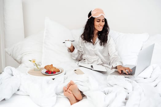 A young lady of Arab appearance starts her working day at the hotel with breakfast in bed. Girl holding a cup of coffee uses a laptop, sitting on a white bed. Best workspace is coziness and comfort