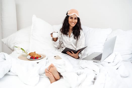 Pretty young woman of Spanish woke up in the morning in bed and starts working day. Croissant and berries a cup of coffee and a notebook in hand - organize the workspace combining with rest