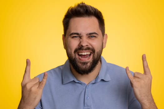 Overjoyed caucasian man showing rock n roll gesture by hands, cool sign, shouting yeah with crazy expression, dancing, emotionally rejoicing in success. Young happy guy isolated on yellow background