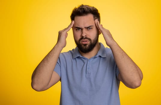 Sick ill caucasian bearded young man rubbing temples to cure headache problem, suffering from tension, migaine, stress, grimacing in pain, high blood pressure isolated alone on yellow wall background