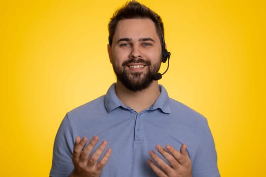 Smiling young man wearing headset, freelance worker, call center or support service operator helpline, having talk with client or colleague, communication support. Guy isolated on yellow background