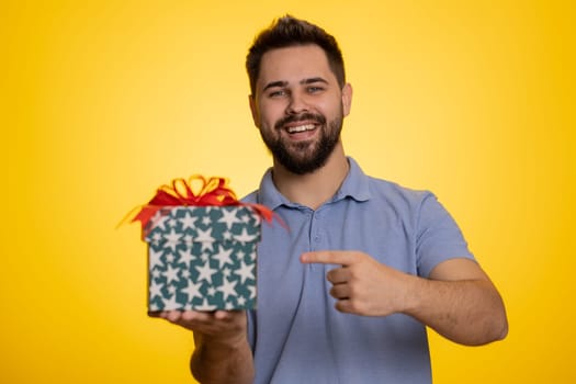 Positive smiling young man presenting birthday gift box stretches out hands, offer wrapped present career bonus, celebrating party, sale. Bearded Caucasian guy isolated on yellow studio background