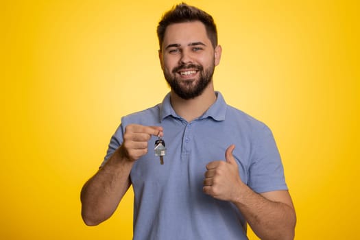 Happy young man real estate agent lifting hand showing the keys of new home house apartment, buying or renting property, mortgage loan. Handsome guy isolated alone on yellow studio background indoors