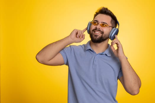 Young happy man listening music via headphones and dancing disco fooling around having fun expressive gesticulating hands, celebrating. Handsome winner guy isolated on yellow studio background indoors