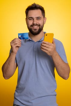 Sincere man customer using credit bank card and smartphone while transferring money, purchases online shopping, payment. Finance and internet. Happy guy isolated alone on yellow background. Vertical