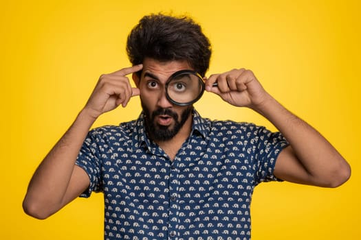 Investigator researcher scientist man holding magnifying glass near face, looking into camera with big zoomed funny eyes, searching, analysing. Handsome indian guy isolated on yellow studio background