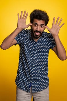 Comical funny young indian man making playful silly facial expressions and grimacing, fooling around, showing tongue. Handsome hindu crazy guy bullying isolated on studio yellow background indoors