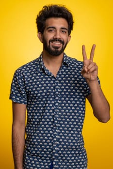 Young indian man showing victory sign, hoping for success and win, doing peace gesture, smiling with kind optimistic expression. Handsome bearded hindu guy isolated alone on yellow studio background