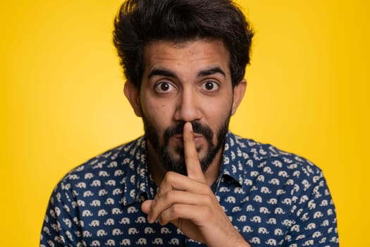 Shh be quiet please. Young indian man presses index finger to lips makes silence gesture sign do not tells secret, stop gossip. Handsome bearded hindu guy isolated alone on yellow studio background