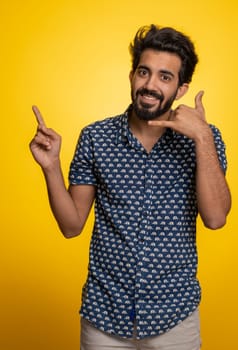Call us, here is contact number. Handsome bearded indian man in shirt looking at camera doing phone gesture like says hey you call me back conversation. Hindu guy isolated on yellow background, indoor