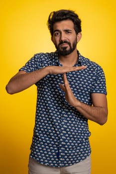 Tired serious upset young indian man showing time out gesture, limit or stop sign, no pressure, I need more time, relax, break, deadline. Handsome hindu guy isolated on studio yellow background indoor