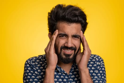 Handsome indian bearded man in shirt rubbing temples to cure headache problem, suffering from tension, migaine, stress, grimacing in pain, high blood pressure isolated alone on yellow wall background