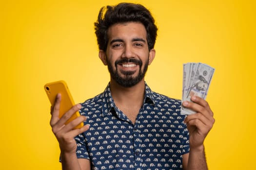 Indian man looking smartphone display sincerely rejoicing win, receiving money dollar cash banknotes, success lottery jackpot luck, income salary wage. Hindu guy isolated on studio yellow background