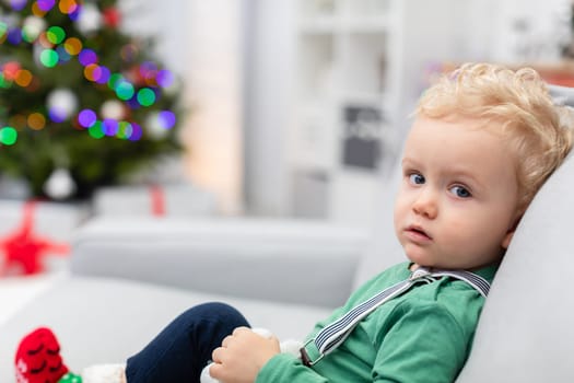 Close-up of a little boy sitting on a gray couch. The child is wearing a bright green sweatshirt and striped suspenders. The boy is wearing red Christmas socks. Blurred background. In the background is a Christmas tree with colorful lights.