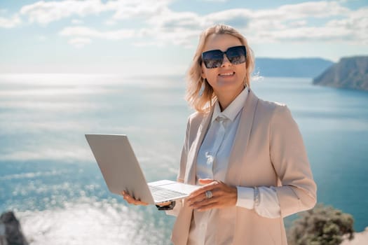 Freelance women sea. She is working on the computer. Good looking middle aged woman typing on a laptop keyboard outdoors with a beautiful sea view. The concept of remote work