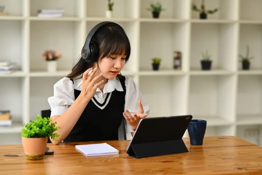 Pretty asian female student wearing headphones learning online, watching webinar class on laptop.