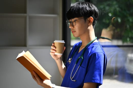 Medical student man wearing blue scrubs reading book, preparing for university exams. Medical internship concept.