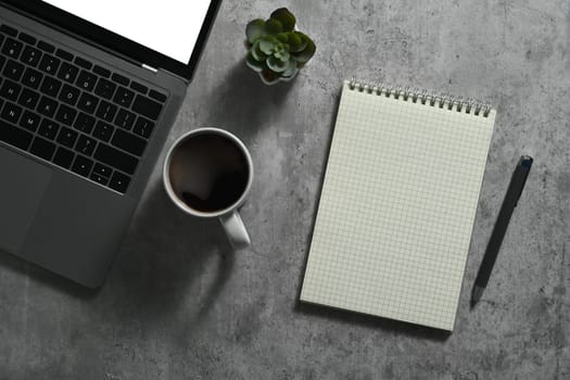 Laptop with blank screen, cup of coffee and notebook on concrete stone office desk. Top view with copy space.