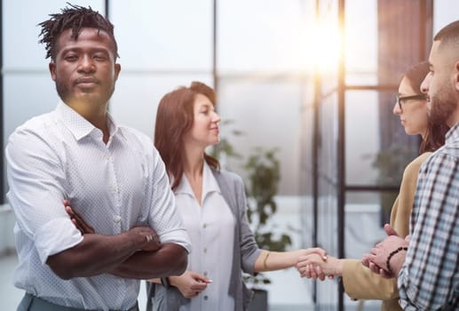 African American man posing for the camera with his arms crossed in front of his colleagues shaking hands in the office