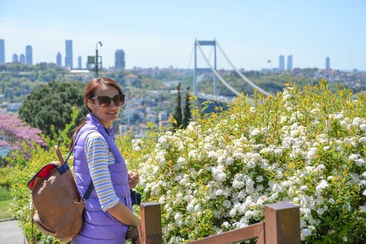 Young beautiful woman tourist with a backpack looks at the camera on the background of the urban landscape overlooking the Fatih Sultan Mehmet Bridge. Back view. Otagtepe Park.