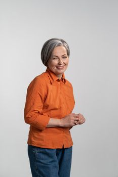 Middle aged grey haired pretty woman smile gentle looking at camera with touching finger tips wearing orange shirt and denim jeans isolated on white background. Human emotions concept.